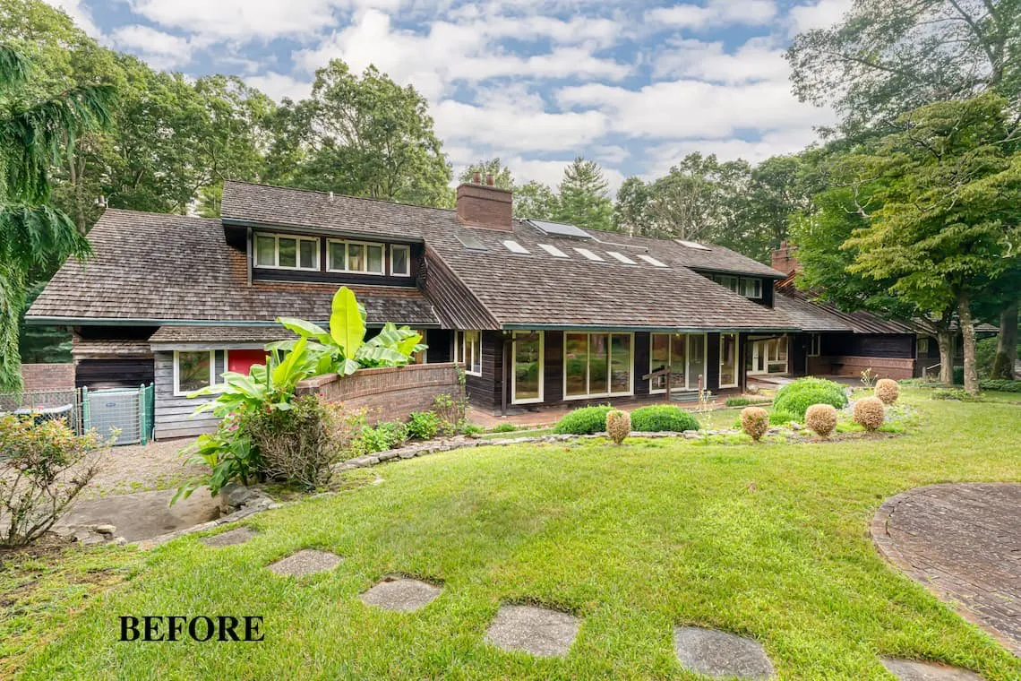 Exterior of a traditional home with dark wood siding, a shingled roof, large windows, and a grassy yard surrounded by trees.