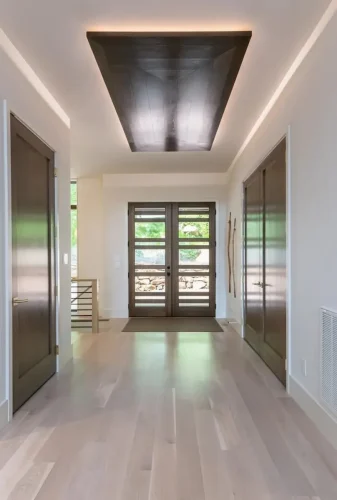Modern entry hallway with wood double doors, light hardwood flooring, dark interior doors, and an illuminated wood ceiling feature.