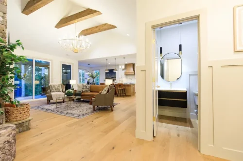 Open living area with light wood floors, exposed ceiling beams, and seating beside large windows, with views into the kitchen and a small bathroom.
