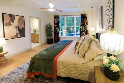 Bedroom with layered cream and green bedding, bedside lamps, patterned curtains, and a large window overlooking trees.