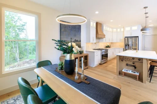 Dining area with green chairs and a circular pendant light beside a white kitchen with a wood island and stainless steel appliances.