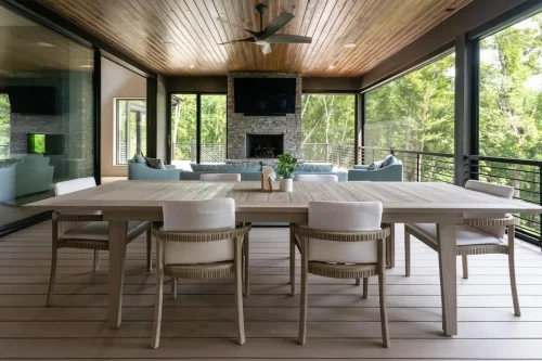 Screened outdoor dining and living area with a wood-paneled ceiling, stone fireplace, and views of the surrounding woods.