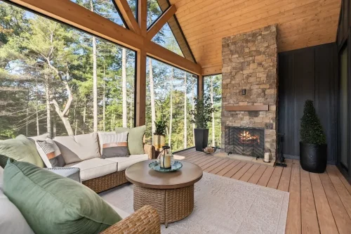 Screened porch overlooking woods, with a vaulted wood ceiling, exposed beams, cushioned seating, and a stone fireplace.