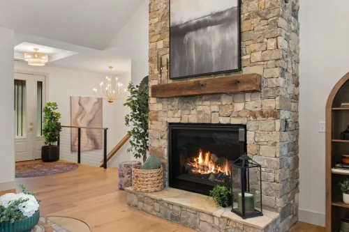 Floor-to-ceiling stone fireplace with a wood mantel and artwork above a burning fire, surrounded by light wood flooring.