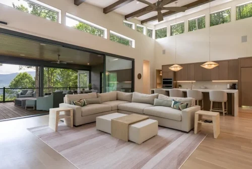 Open great room with vaulted ceiling, exposed wood beams, large windows, modern kitchen, and sliding doors opening to a covered deck.