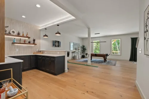 Open recreation area with a dark-cabinet bar, floating shelves, pendant lights, and a pool table near large windows.