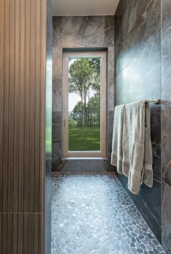 Walk-in shower with dark stone tile walls, pebble tile flooring, and a tall window overlooking the wooded landscape.