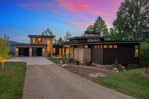 Contemporary mountain home at dusk with low rooflines, stone accents, warm exterior lighting, and an attached two-car garage.