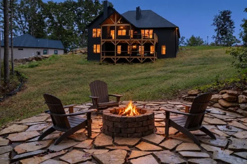 Adirondack chairs surround a lit stone fire pit on a flagstone patio, with the home’s illuminated rear porches beyond the lawn.