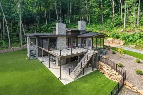Rear aerial view of a contemporary mountain home with stone chimneys, expansive decks, large windows, and landscaped outdoor areas.