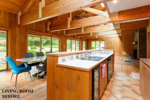 Living area before renovation featuring exposed wood beams, wood-paneled walls, vaulted ceilings, and an open kitchen.