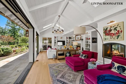 Renovated living room with white vaulted ceilings, pink upholstered seating, fireplaces, built-in shelving, and large sliding glass doors opening to a landscaped patio.