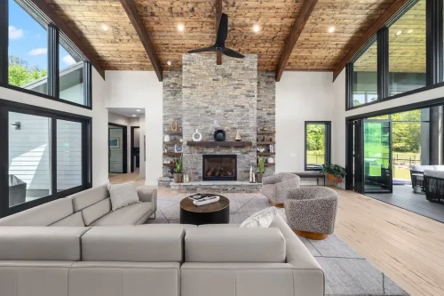 Open living room with a floor-to-ceiling stone fireplace, vaulted wood ceiling, exposed beams, and large black-framed windows.