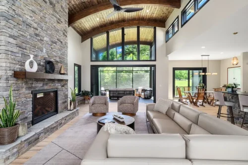 Spacious living area with a stone fireplace, curved wood ceiling, large windows, and an open connection to the dining and kitchen areas.