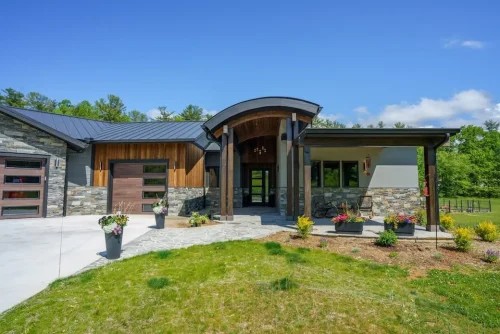 Contemporary mountain home with stone and wood siding, a curved covered entry, dark metal roof, and landscaped front patio.