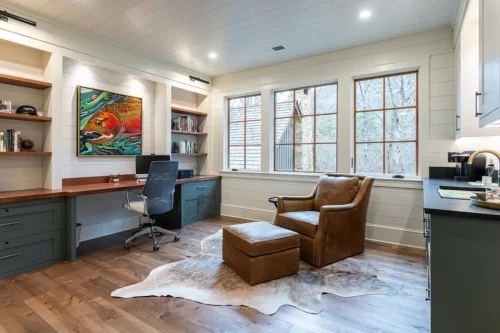 Home office with built-in desk and shelving, green cabinetry, large windows, hardwood floors, and a leather chair.