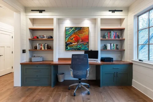 Built-in home office with wood desktop, green cabinetry, open shelving, and colorful artwork.