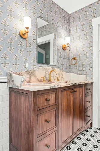 Angled view of a wood vanity with marble countertop, brass fixtures, floral wallpaper, and patterned tile flooring.