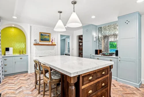 Large marble-topped wood island surrounded by light blue cabinetry, pendant lighting, and herringbone brick flooring.