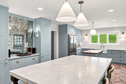 Wide view of the kitchen featuring a marble-topped island, light blue cabinetry, stainless steel appliances, and herringbone brick flooring.
