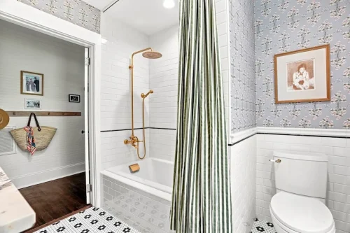 Bathroom with a white tiled tub and shower, floral wallpaper, brass fixtures, and patterned tile flooring.