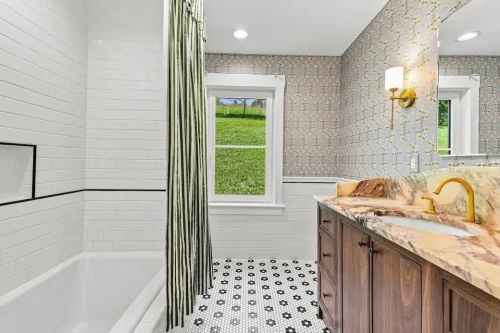 Bathroom with a wood vanity, marble countertop, floral wallpaper, brass fixtures, and white tile.