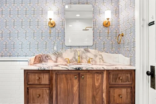 Wood vanity with a marble countertop, floral wallpaper, brass fixtures, and wall sconces.