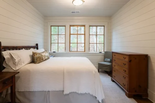 Bedroom with white shiplap walls, hardwood floors, wood furniture, and three large windows overlooking the woods.