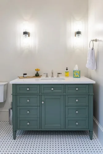 Bathroom with a green vanity, white countertop, wall sconces, and black-and-white tile flooring.