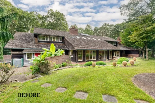 Exterior of a traditional home with dark wood siding, a shingled roof, large windows, and a grassy yard surrounded by trees.
