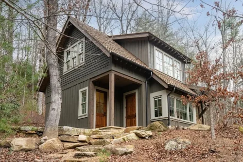 Exterior of a dark mountain carriage house with wood accents, stone landscaping, and surrounding trees.
