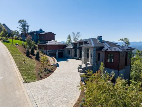 Angled aerial view of the home featuring a large paved driveway, stone columns, dark metal roof, and mountain setting.