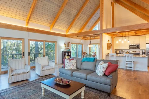 Open living room and kitchen with exposed wood beams, vaulted ceilings, and large windows overlooking the wooded setting.