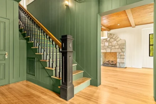 Interior entry with green paneled walls, wood staircase, black railing, hardwood floors, and a stone fireplace visible beyond.