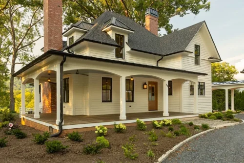 White two-story home with wraparound covered porch, brick chimneys, black-framed windows, and landscaped front yard.