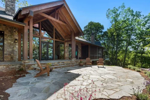Rustic mountain home with timber beams, stone facade, and large windows opening onto a spacious flagstone patio with Adirondack chairs, surrounded by trees.