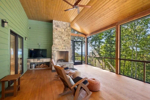 Inviting screened-in porch with vaulted wood ceiling, stone fireplace, mounted TV, and rocking chairs overlooking the forest and distant views.