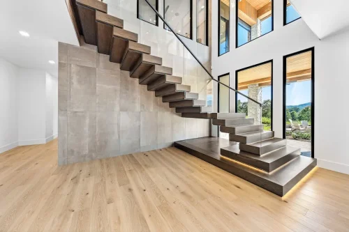 Floating wood staircase with glass railing and tall windows filling the space with natural light.