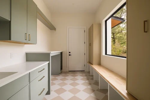 Laundry and utility room featuring sage green cabinetry, white countertops, brass hardware, checkered tile flooring, and a built-in wood bench.