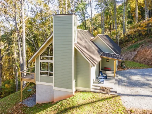 Exterior view of a green mountain home with a metal roof, tall chimney, large windows, and elevated deck.