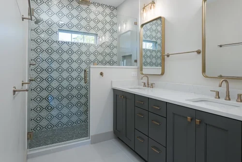 Bathroom with a double vanity, brass fixtures, and a glass-enclosed shower with geometric patterned tile.