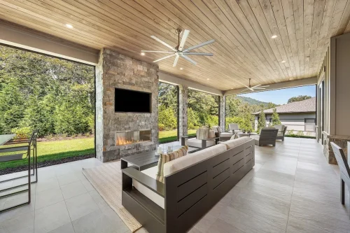 Covered patio with stone fireplace, TV, and lounge seating overlooking trees.