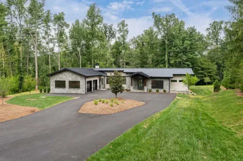 Contemporary single-story home with stone and light-colored siding, black metal roof, circular driveway, and landscaped lawn surrounded by trees.