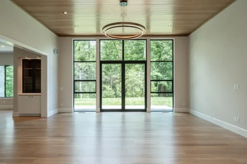 Spacious living area featuring a wood-paneled ceiling, circular pendant light, light hardwood flooring, and floor-to-ceiling black-framed windows overlooking the backyard.