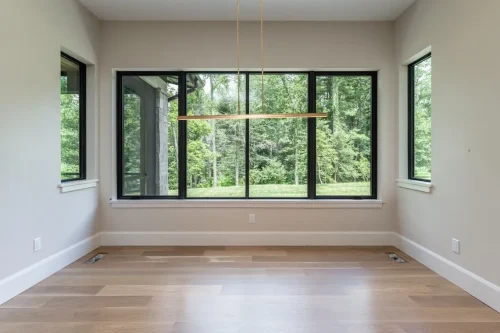 Bright dining area featuring large black-framed windows overlooking the wooded backyard, light hardwood floors, and a linear pendant light.