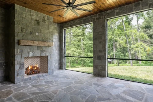 Covered outdoor living space with stone walls and flooring, a wood-paneled ceiling, ceiling fan, and stone fireplace overlooking the backyard.