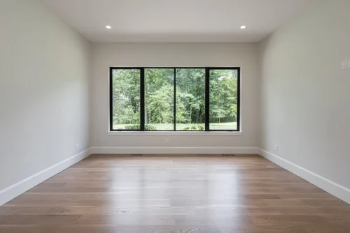 Unfurnished bedroom with light hardwood flooring, white walls, and a large black-framed window overlooking the trees.