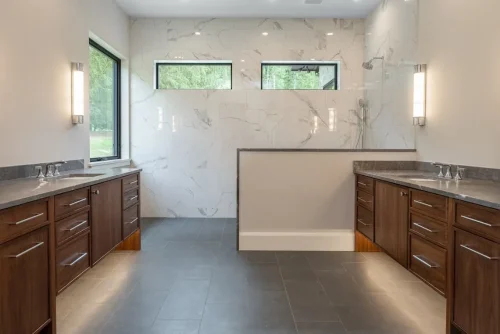 Spacious bathroom featuring two dark wood vanities, gray countertops, marble-look tile walls, and a walk-in shower.