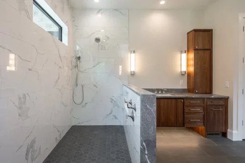 Large walk-in shower with marble-look tile walls, dark hexagonal floor tiles, and a half-wall separating the shower from a wood vanity.