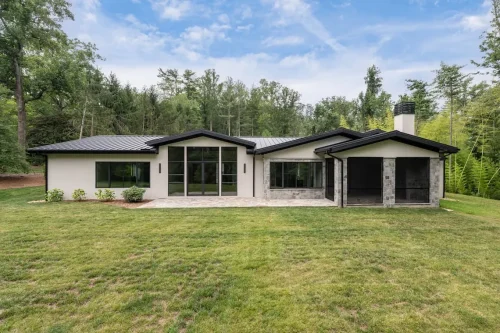 Rear view of a contemporary single-story home with black metal rooflines, large black-framed windows, stone accents, and a screened porch overlooking the lawn.